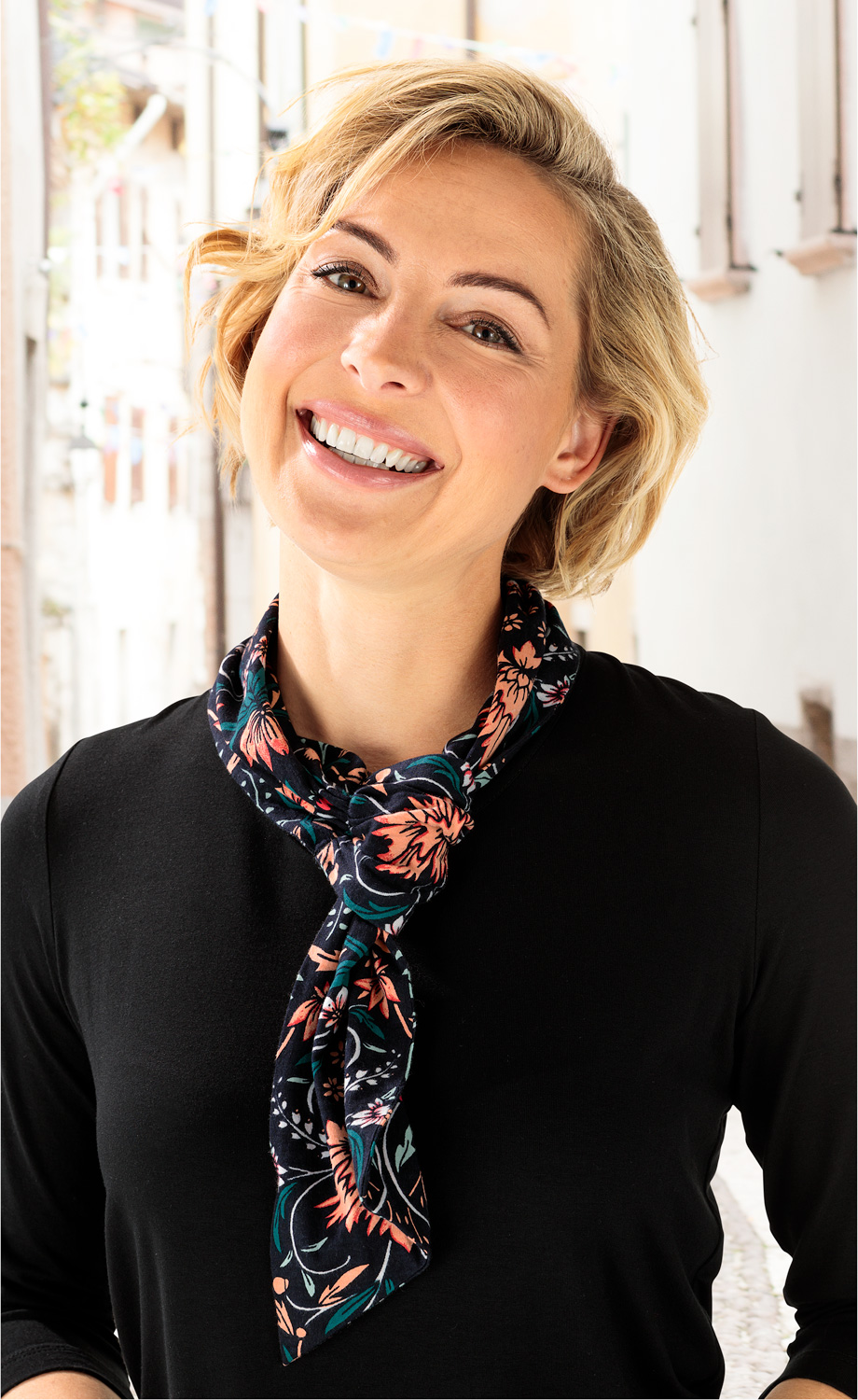 Frau mit kurzem blondem Haar, lächelnd, trägt schwarzes Baumwoll-Mix-Kleid und dunkelblauen, floral gemusterten Schal, im hellen urbanen Hintergrund mit Gebäuden und Fenstern.