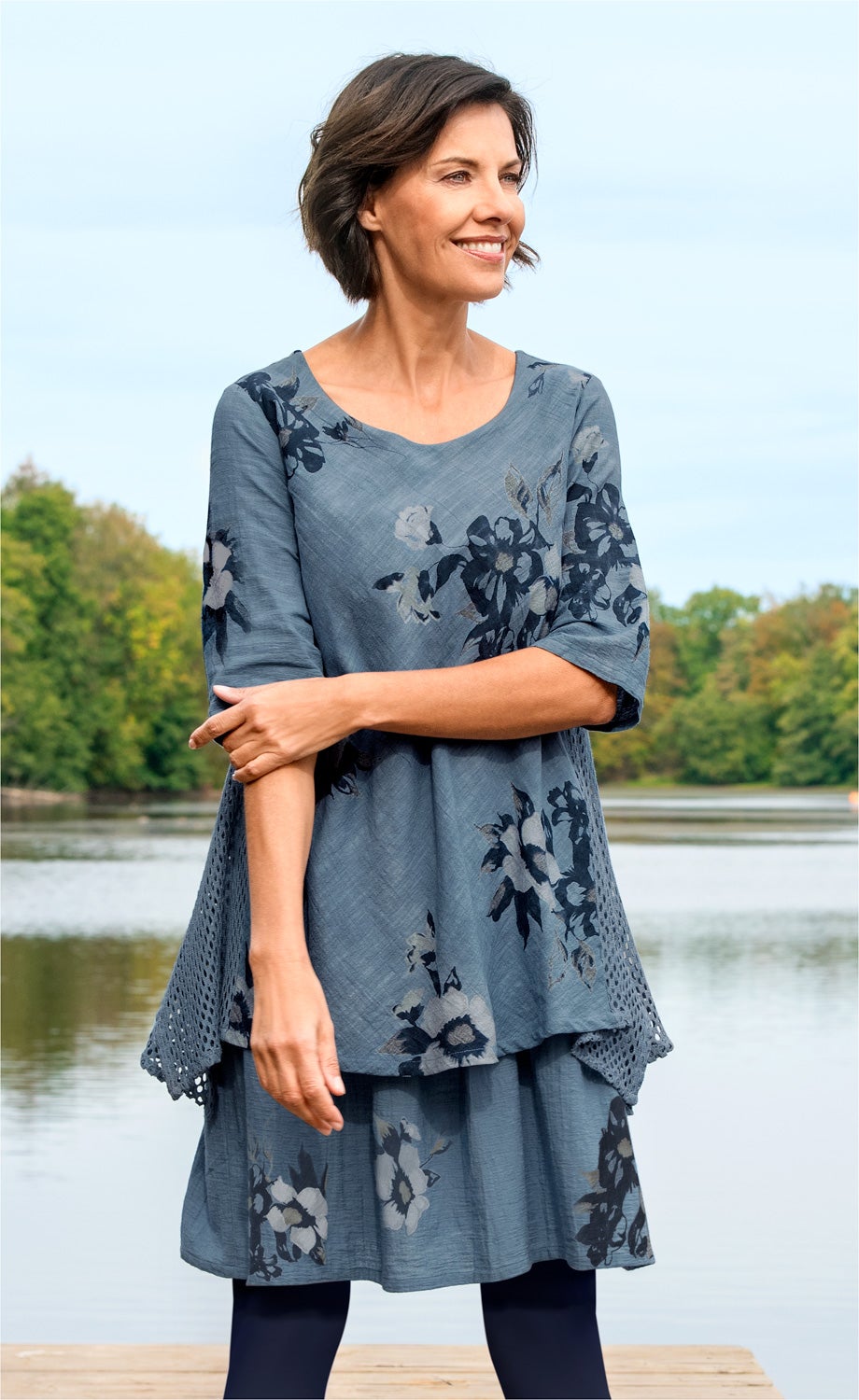 Frau mit kurzem braunem Haar trägt blau-graues Baumwoll-Kleid mit floralem Muster und 3/4-Ärmeln, steht an einem Seeufer mit bewaldetem Hintergrund, lächelt und schaut seitlich.