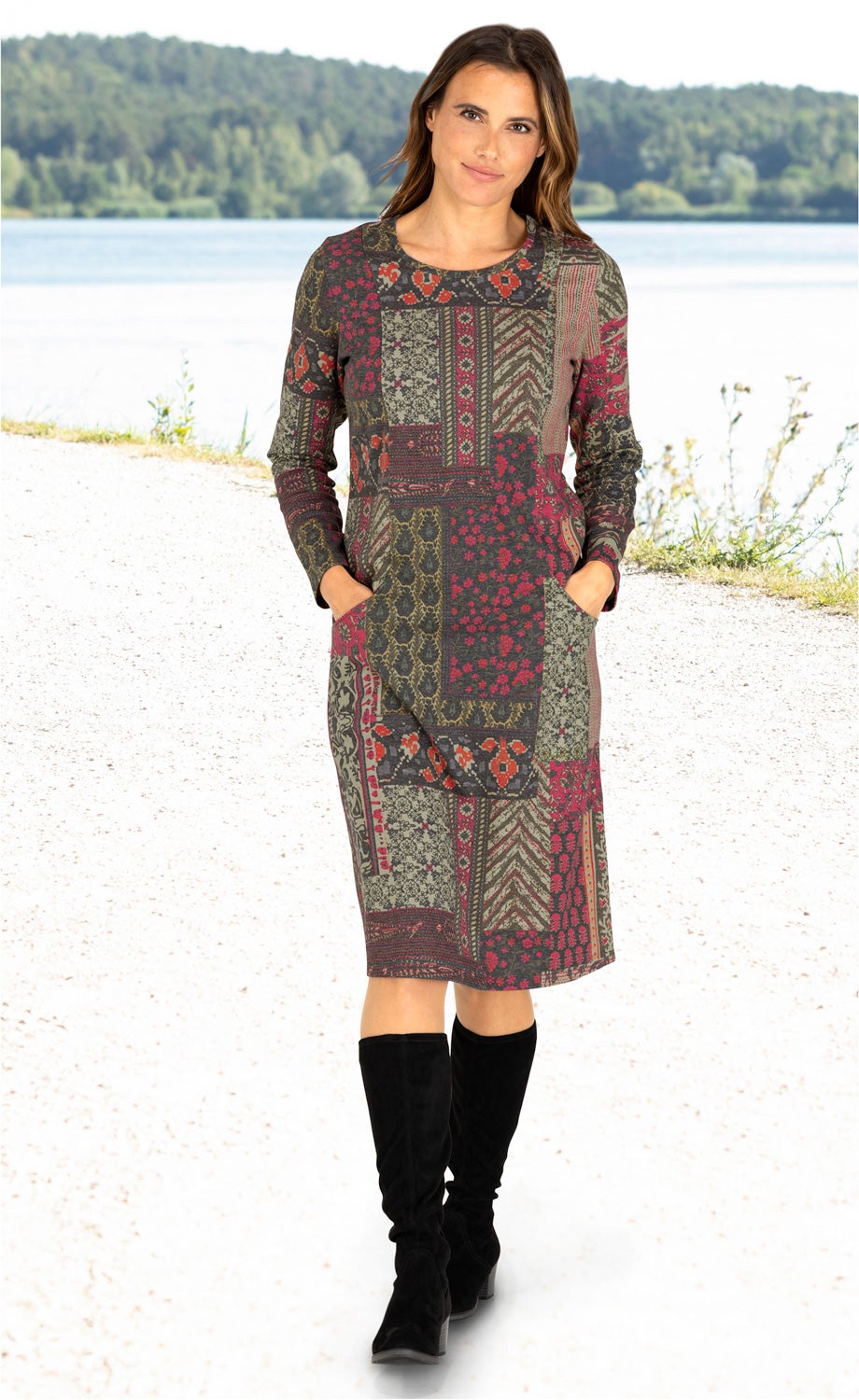 Frau trägt knielanges Baumwoll-Mix-Kleid mit langen Ärmeln und buntem Patchwork-Muster in Rot, Grün und Grau, zwei Taschen vorne, dazu schwarze kniehohe Stiefel, stehender Porträt vor See und Wald im Hintergrund