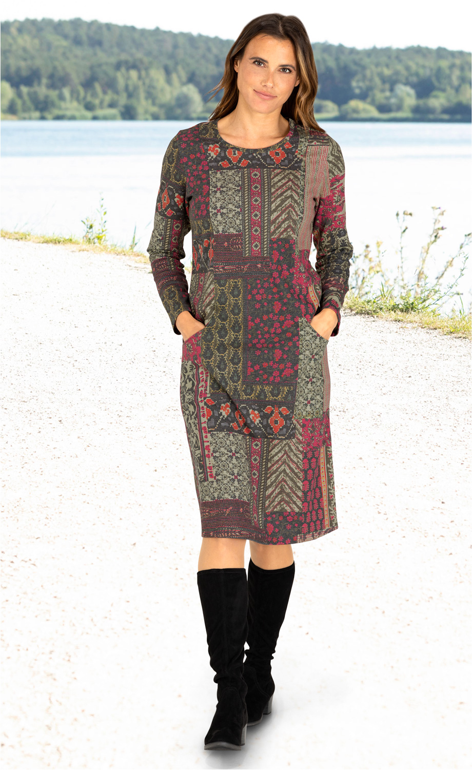 Frau trägt knielanges Baumwoll-Mix-Kleid mit langen Ärmeln und buntem Patchwork-Muster in Rot, Grün und Grau, zwei Taschen vorne, dazu schwarze kniehohe Stiefel, stehender Porträt vor See und Wald im Hintergrund