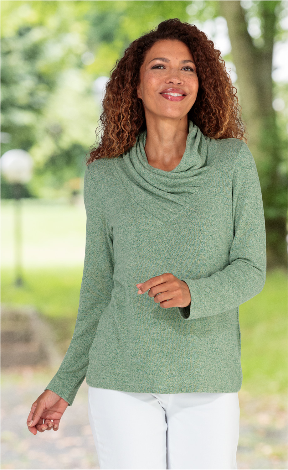 Frau trägt grünes Viskose-Mix-Shirt mit schmalem Stehkragen, lange Ärmel, lächelnd, stehendes Porträt, weißer Hose, unscharfer grüner Parkhintergrund an sonnigem Tag