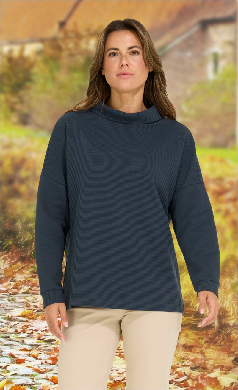 Frau trägt dunkelblaues langärmeliges Baumwoll-Shirt mit Stehkragen, beigefarbene Hose, herbstlicher Hintergrund mit Laub und unscharfen Häusern