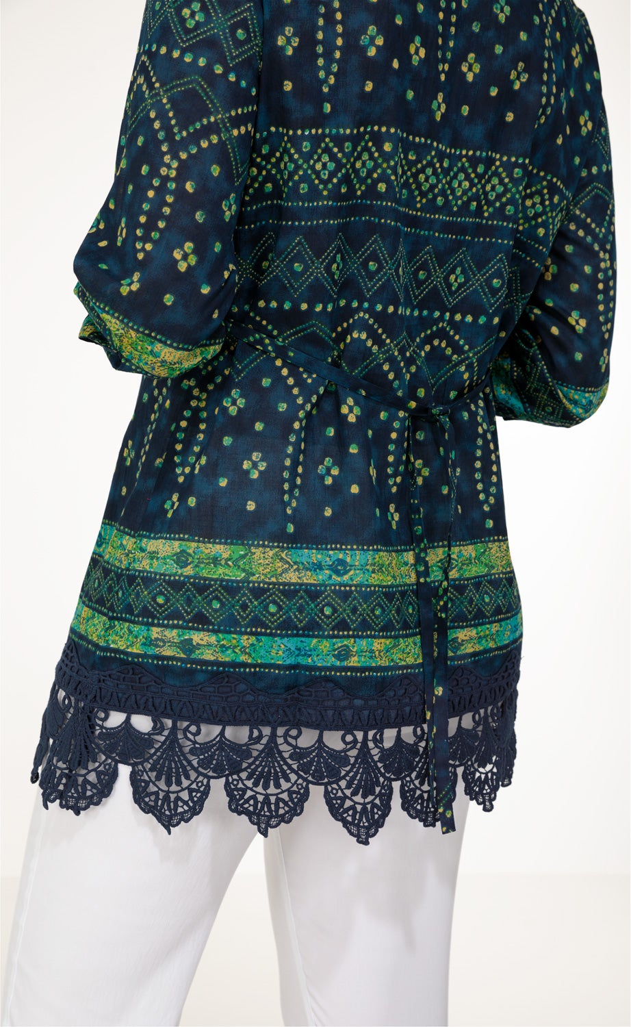 Dunkelblaue Baumwoll-Bluse mit grünen und gelben Ethno-Mustern, Bindegürtel in Taillenhöhe, langärmlig, mit gehäkeltem Spitzenabschluss am Saum, getragen zu weißer Hose, Ansicht von hinten.