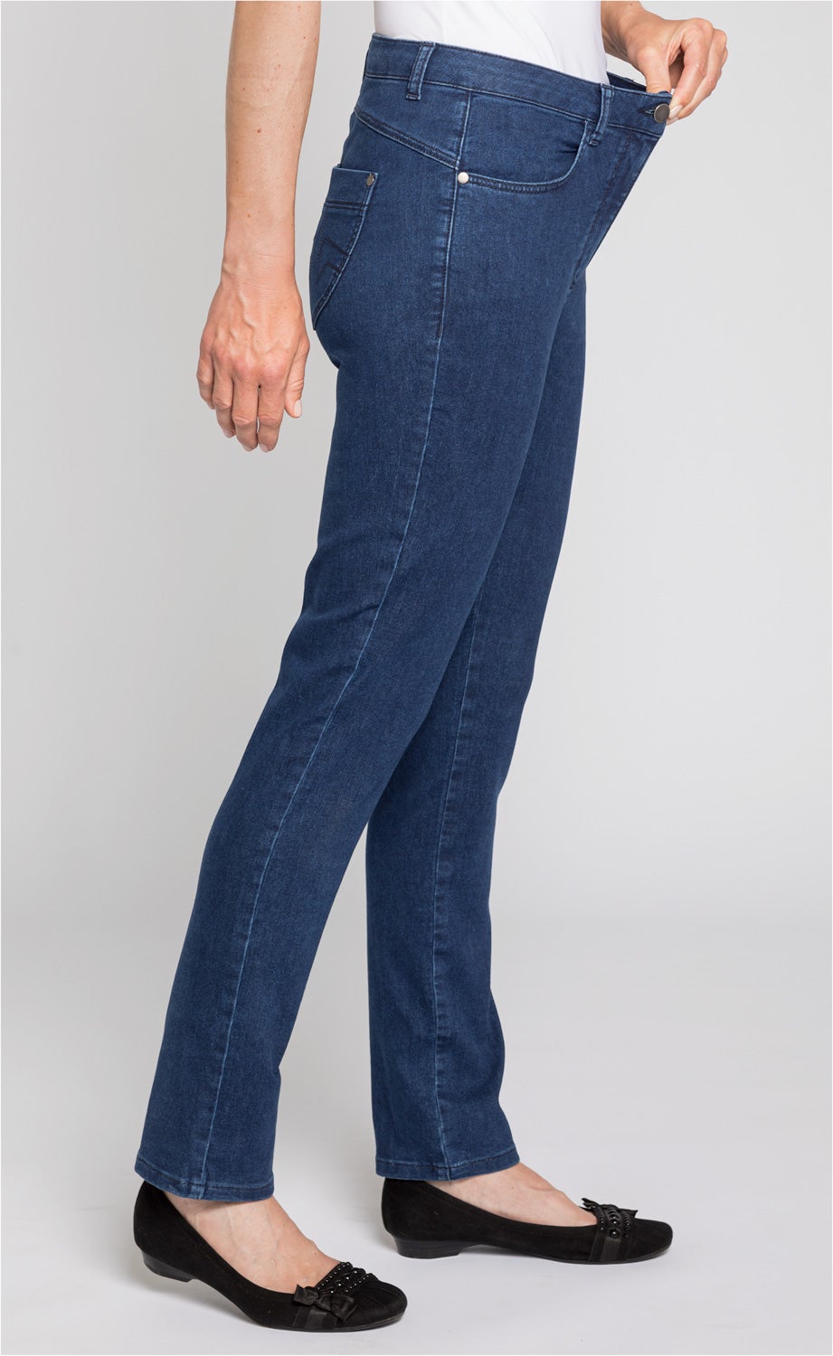 Seitliche Ansicht einer dunkelblauen Baumwoll-Mix-Hose, die an der Taille von einer Person mit weißem Oberteil leicht ausgezogen wird, kombiniert mit schwarzen flachen Schuhen mit Schleifenapplikation.