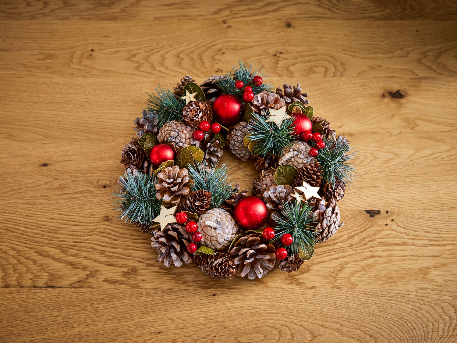 runder Deko-Kranz aus Tannenzapfen, grünen Kiefernzweigen, roten Weihnachtskugeln, roten Beeren und kleinen Holzsternen auf hellem Holztisch