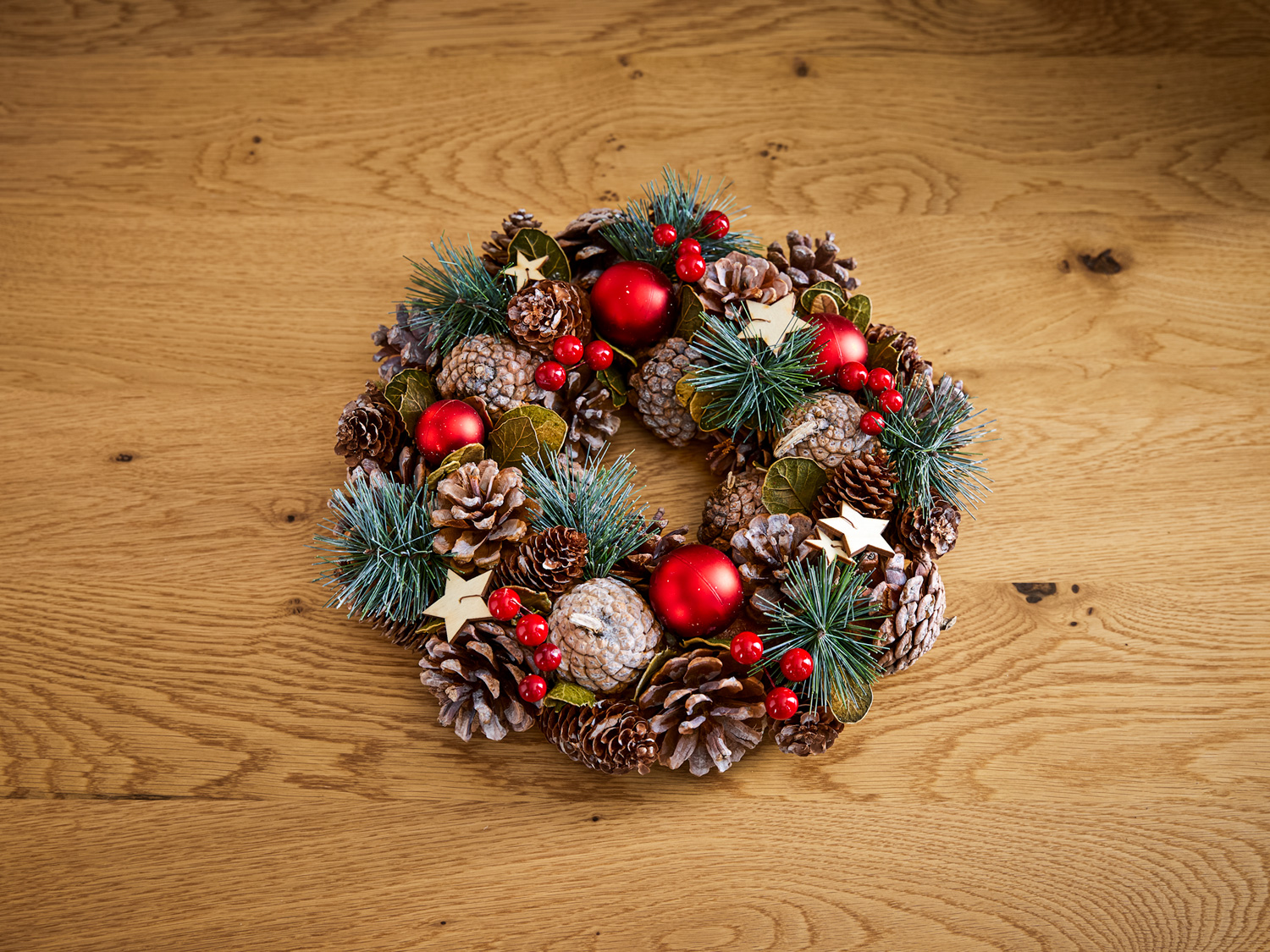 runder Deko-Kranz aus Tannenzapfen, grünen Kiefernzweigen, roten Weihnachtskugeln, roten Beeren und kleinen Holzsternen auf hellem Holztisch