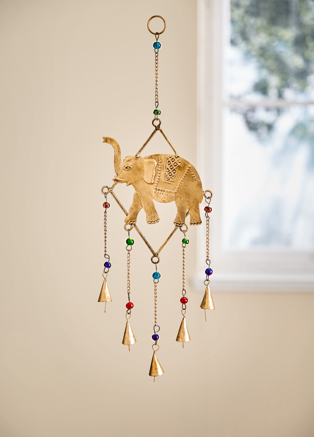 Metall-Mobile 48cm mit einem goldfarbenen Elefanten in der Mitte, verziert mit kleinen bunten Perlen, hängend an einer Kette mit mehreren kleinen Glocken, vor hellem Hintergrund und Fenster mit unscharfen Baumblättern.