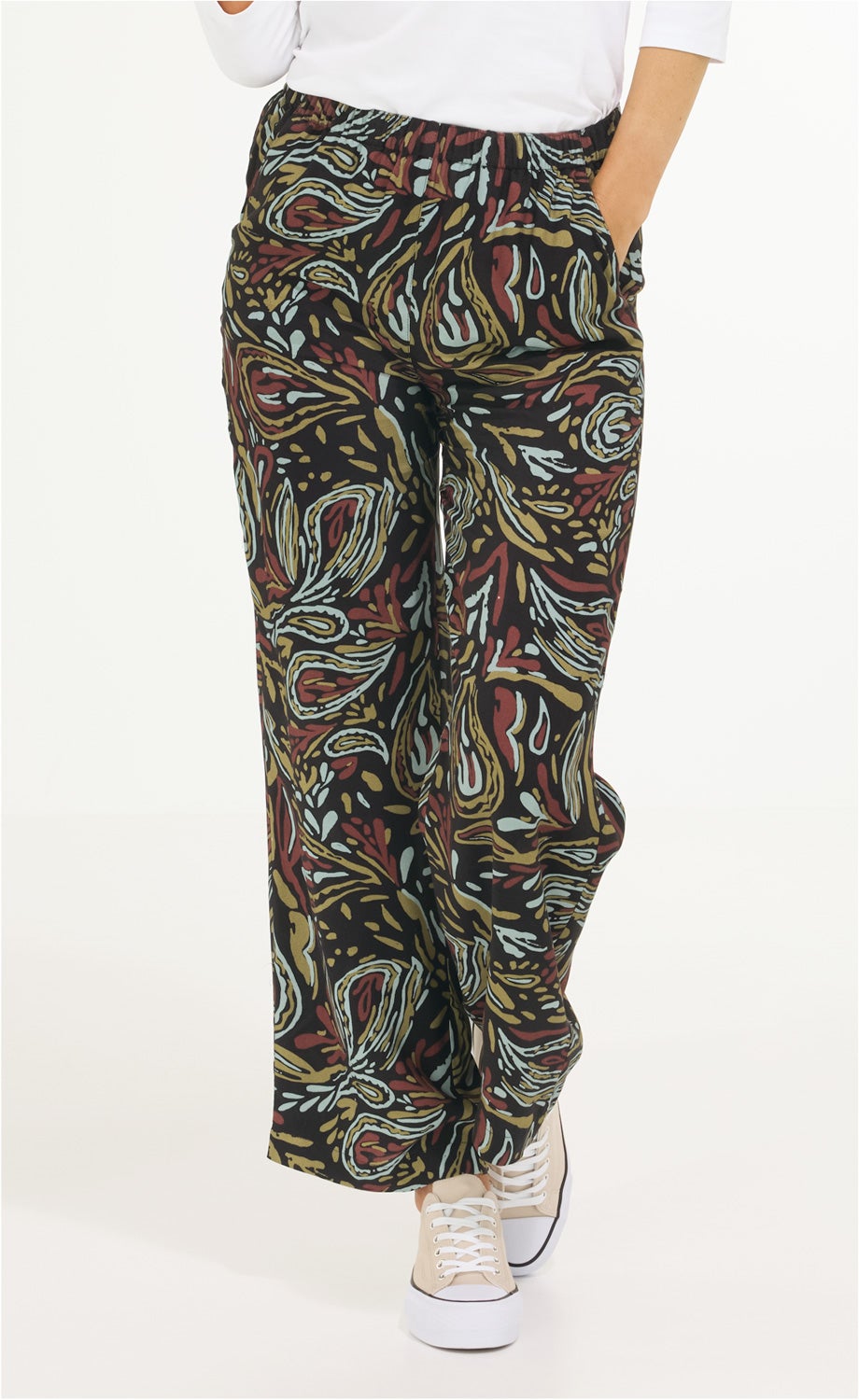 Damen trägt Viskose-Hose mit weitem Bein, abstraktem Muster in Schwarz, Braun, Grün und Hellblau, elastischem Bund, kombiniert mit weißem Oberteil und beigen Sneakern mit weißen Schnürsenkeln