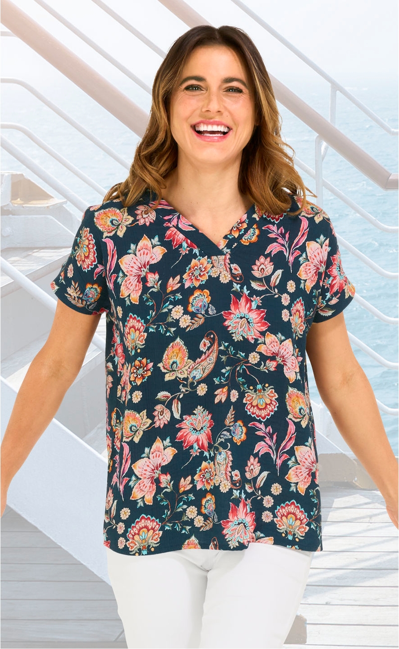Frau trägt kurzärmelige Baumwoll-Bluse mit dunkelblauem Grund und buntem floralen Muster, lächelt, sitzt vor Treppengeländer mit Blick aufs Meer, weiße Hose.