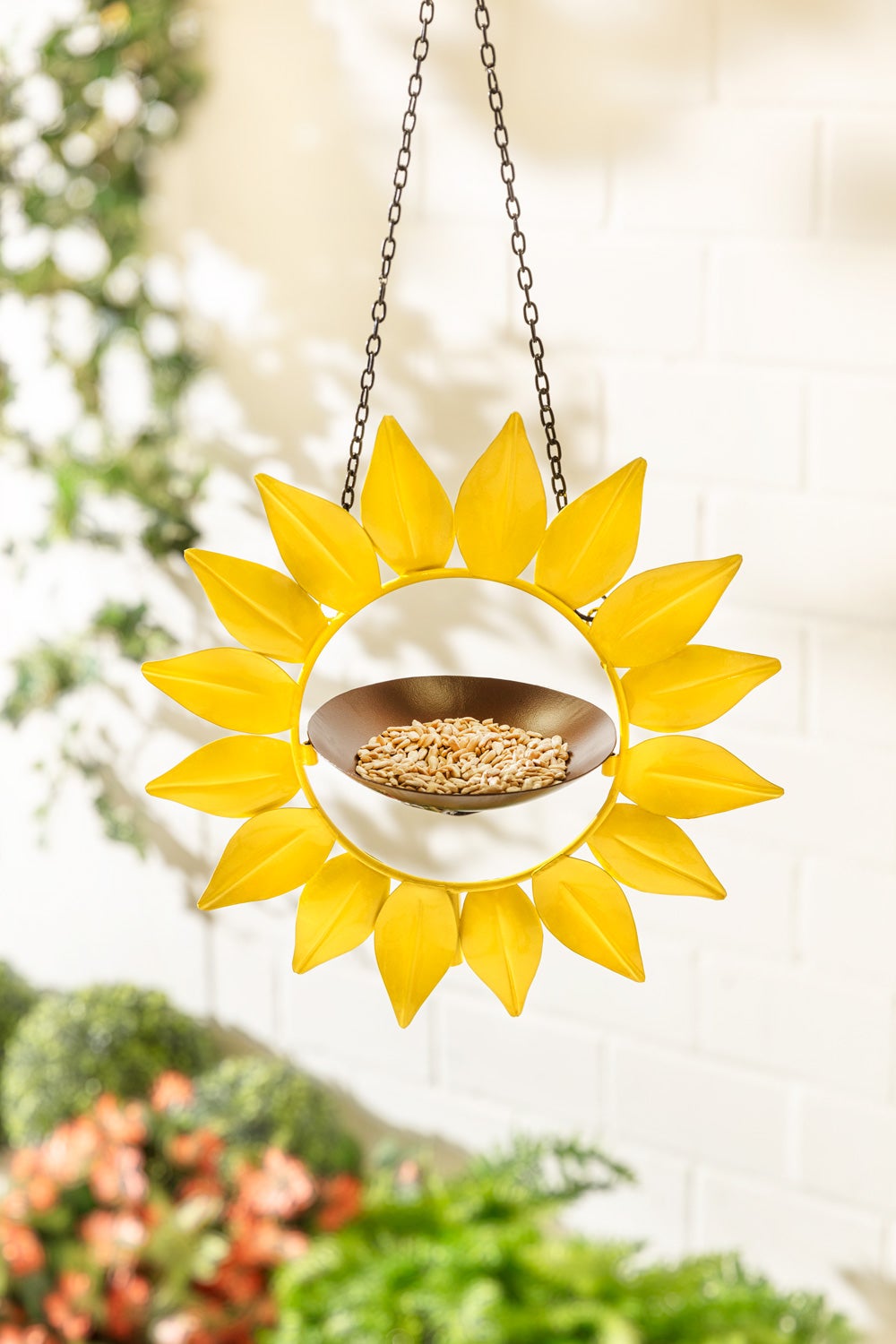 Metall-Futterstation in Sonnenblumenform, gelbe Metallblätter, hängend an Kette, braune Futterschale mit Vogelfutter, unscharfer Hintergrund mit weißen Wand und grünen Pflanzen.
