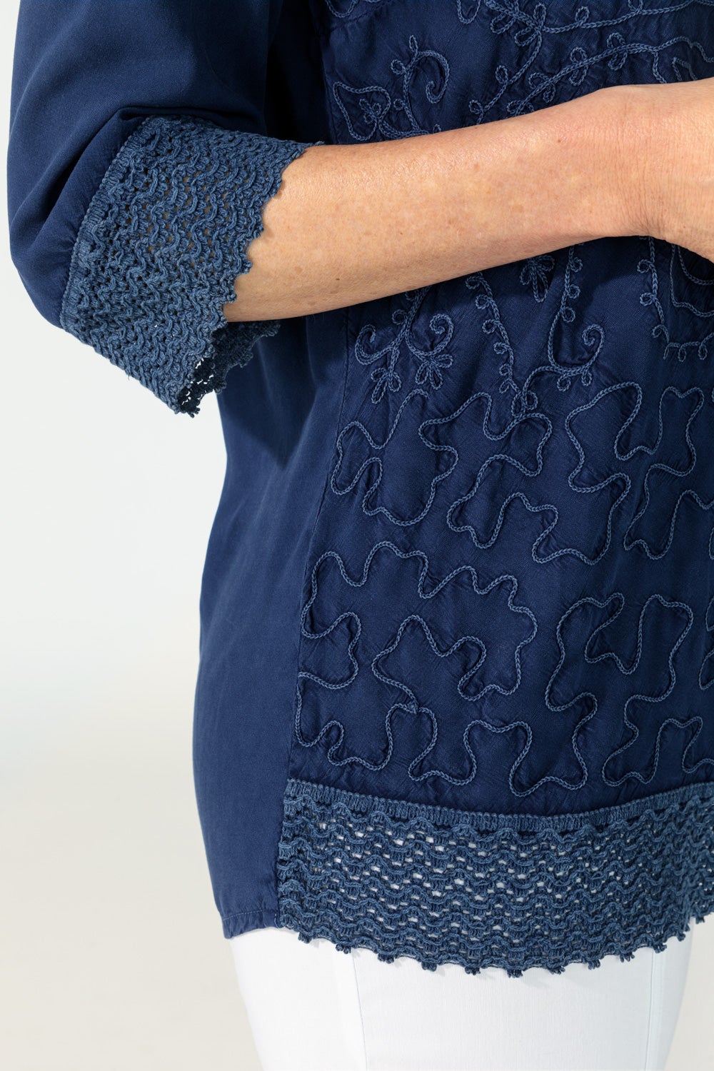 Detailaufnahme einer dunkelblauen Viskose-Bluse mit floraler Stickerei, gehäkeltem Abschluss am Ärmel und Saum, getragen von einer Person mit hellem Hautteint und weißer Hose