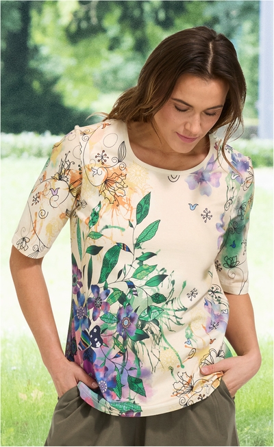 Frau trägt cremefarbenes Baumwoll-Shirt mit buntem Blumen- und Blätterdruck, Halbarm, Rundhalsausschnitt, Hände in olivgrüner Hose in den Taschen, stehend vor unscharf-grünem Hintergrund