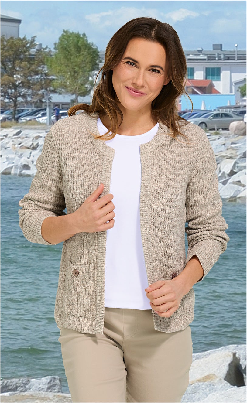 Frau trägt beige Baumwoll-Strickjacke mit zwei Taschen und Knöpfen, weißes T-Shirt und beige Hose, steht am Wasser mit Felsen und Häusern im Hintergrund unter blauem Himmel
