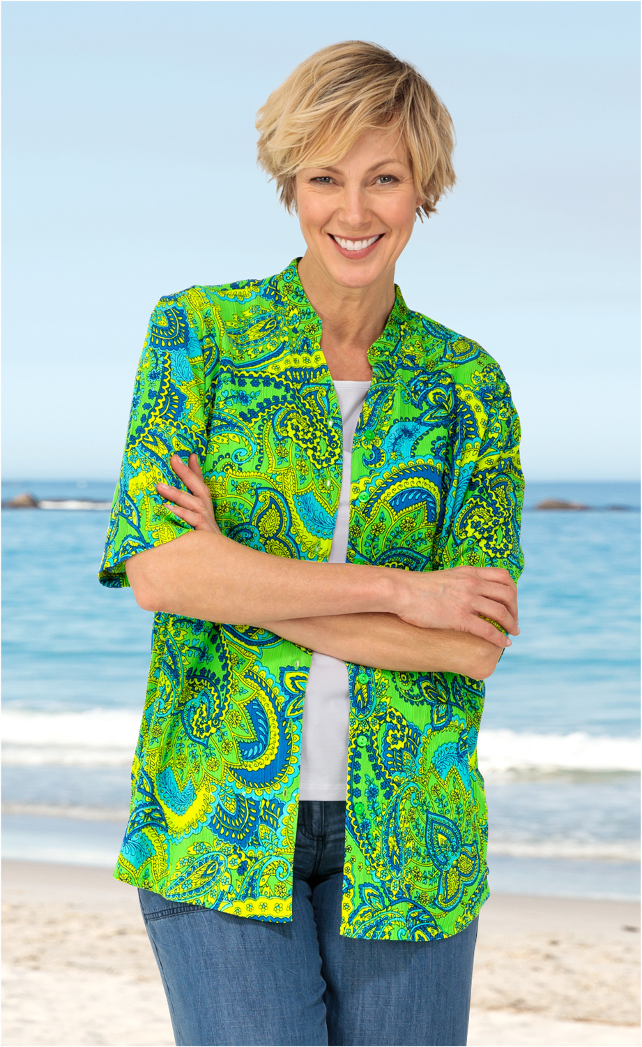 Frau am Strand trägt grüne, blau-gelbe Viskose-Bluse mit Paisley-Muster, weiße Shirt darunter, blaue Jeans, lächelt, Arme verschränkt, heller Himmel und Meer mit Wellen im Hintergrund