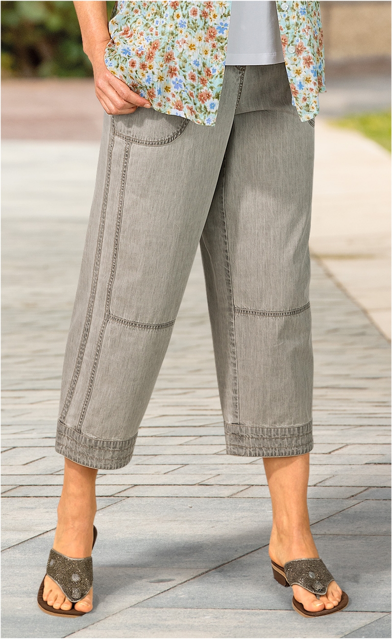 Damen trägt graue, weite Baumwoll-Hose mit aufgesetzten Taschen und abgesteppten Details, kombiniert mit gemusterter Bluse und Sandalen mit Absatz.