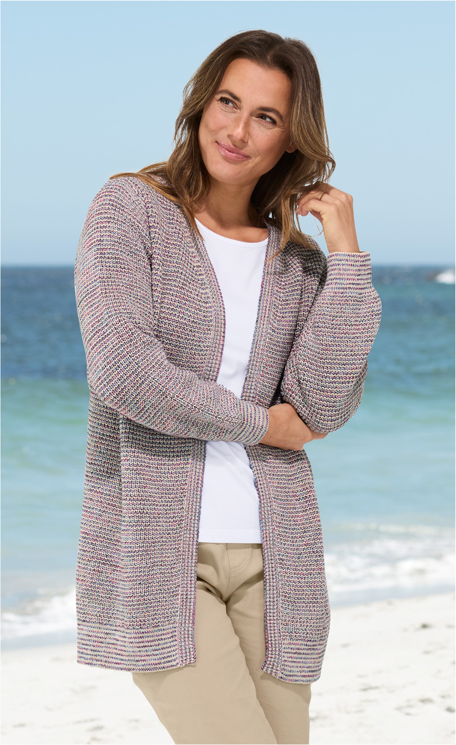 Frau trägt leichte mehrfarbige Baumwoll-Strickjacke mit offenem Schnitt, weiße Bluse, helle Hose, steht am Strand mit Meer im Hintergrund, lächelnd, eine Hand leicht am Haar, helle Tageslichtaufnahme