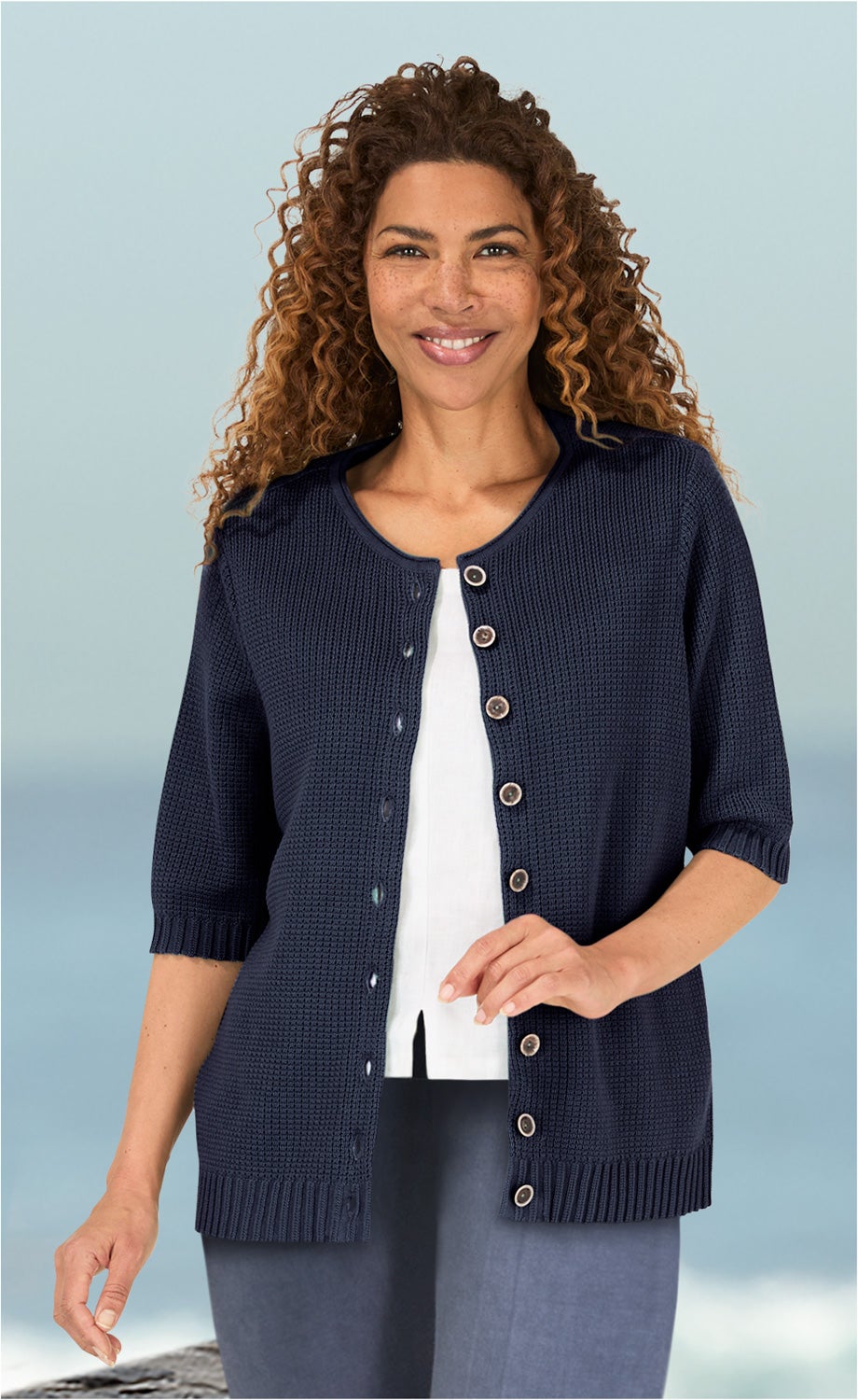 Frau mit lockigem braunem Haar trägt dunkelblaue Baumwoll-Strickjacke mit kurzen Ärmeln und Knopfleiste, weiße Bluse und graue Hose, vor blauem Hintergrund mit Wasser und Felsen sichtbar.