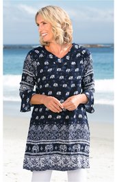 Dunkelblaues Damenoberteil mit weißen Elefanten- und geometrischen Streifenmustern, V-Ausschnitt, 3/4-Ärmel mit Schlitz, lange lockere Passform, getragen von stehender Frau am Strand, seitlich gewelltes hellblondes Haar