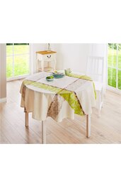 Runder Tisch mit Tischdecke in Creme, Grün und Braun, gemustert mit floralen und einfarbigen Segmenten, Tisch steht in hellem Raum mit Holzfußboden, weißem Stuhl rechts, kleinem Beistelltisch mit Schublade dahinter, Geschirr mit grünem Becher und Tel