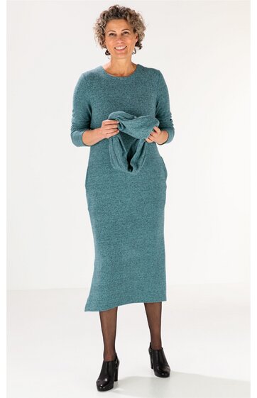 Langärmeliges, tealfarbenes Strickkleid, rundhals, knielang, gerade geschnitten, getragen von stehender Frau mit kurzem lockigem Haar, schwarze Netzstrumpfhose, schwarze Ankle Boots, hält passendes Stricktop in Händen