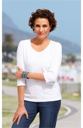 Weißes Langarmshirt mit Rundhalsausschnitt, schlichtes Design, getragen von stehender Frau, kombiniert mit blauen Jeans, Armbänder am linken Handgelenk