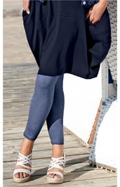 Dunkelblauer Ballonrock mit Taschen, kombiniert mit knapp unter dem Knie endenden grauen Leggings, weiße Sandalen mit mehreren Riemen, stehend auf Holzplanken, Hände in Taschen