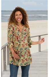 Beige Tunika mit buntem Blumenmuster in Rot, Blau, Grün und Gelb, V-Ausschnitt, lange Ärmel, locker geschnitten, getragen von lächelnder Frau stehend am Strand mit Geländer