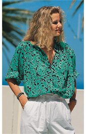 Grüne Bluse mit schwarzem Blumenmuster, V-Ausschnitt, kurze Ärmel mit Bündchen, locker sitzend, getragen von stehender Frau mit seitlich geneigtem Kopf, weiße hohe Hose mit Bund und Taschen