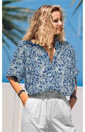 Locker sitzende Bluse in Blau mit weißem Blumenmuster, V-Ausschnitt, kurze Ärmel, getragen von stehender Frau mit offener Pose, kombiniert mit weißen Hosen