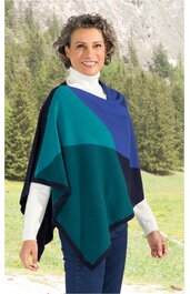 Poncho in verschiedenen Blautönen und Grün, geometrisches Blockmuster, asymmetrischer Schnitt, ohne Verschluss, getragen von stehender Frau mit weißem Rollkragenpullover und Jeans