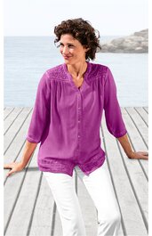 Bluse in magentaviolett, unifarben, mit Knopfleiste vorne, Stehkragen, 3/4-Ärmeln, Spitzenbesatz an Schultern und Saum, getragen von sitzender Frau am Holzsteg mit Blick aufs Wasser