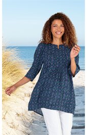 Dunkelblaue Tunika mit geometrischem Muster in Hellblau und Türkis, rundem Ausschnitt mit kleinem Schlitz, 3/4-Ärmeln, locker fallend, getragen von stehender Frau am Strand