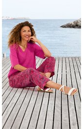 Fuchsiafarbener Pullover mit V-Ausschnitt, rot-pink gemusterte Hose mit floralem Design, weiß-beige Sandaletten, sitzende Frau auf Holzsteg am Wasser, entspannte Pose, gerades Schnittmuster