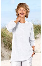 Weißer Pullover ohne Muster, langer Schnitt mit Rundhalsausschnitt, lange Ärmel, getragen von lächelnder stehender Frau mit kurzen blonden Haaren vor Strandkulisse