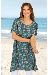 Tunika in dunklem Petrol mit floralem Blumenmuster in Rosa und Hellgrün, kurze Ärmel mit Gummizug, rundhals, tailliert, seitliche Schlitze, getragen von lächelnder Frau am Strand