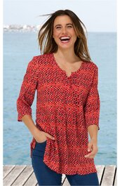 Rotes Langarmshirt mit dunkelblauem geometrischem Muster, V-Ausschnitt mit kleiner Knopfleiste, locker sitzend, getragen von stehender Frau vor Meereskulisse