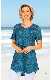 Blaugrünes T-Shirt mit abstraktem Blumenmuster, Rundhalsausschnitt, kurze Ärmel, leicht tailliert, unten mit asymmetrischem Saum, getragen von stehender Frau an Strandkulisse