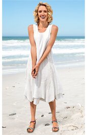 Ärmelloses, weißes Midikleid mit V-Ausschnitt, weitem Rock und strukturiertem Stoff, getragen von stehender Frau mit lockeren Haaren am Strand, Sand unter den Füßen, Sandalen mit bunten Riemen