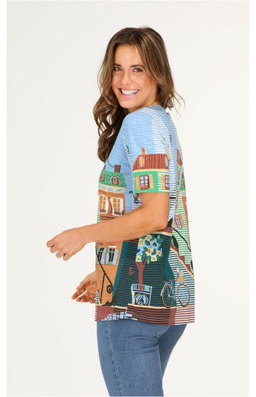Shirt mit hellblauem Oberteil und buntem Dorfmotiv mit Häusern, Bäumen, Fahrrad auf Vorderseite, kurze Ärmel, gerader Schnitt, getragen von seitlich stehender Frau mit lockigen braunen Haaren