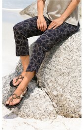 Schwarze, knöchellange Hose mit weißem, geometrischem Punktmuster, gerade geschnitten, getragen von sitzender Person auf hellem Felsen, kombiniert mit beigem Oberteil und schwarzen Sandalen mit goldfarbenen Verzierungen