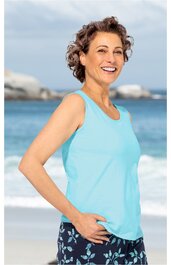 Hellblaues ärmelloses Top mit rundem Ausschnitt, getragen von lachender stehender Frau, dunkelblaue Hose mit hellem Blumenmuster, Strand und Meer im Hintergrund