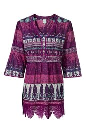 Tunika in dunkelviolett mit weißen und dunkelblauen floralen und geometrischen Mustern, V-Ausschnitt mit Knopfleiste, 3/4-Ärmel mit leichter Glockenform, seitliche Schlitze, unterer Saum mit Spitzenbesatz