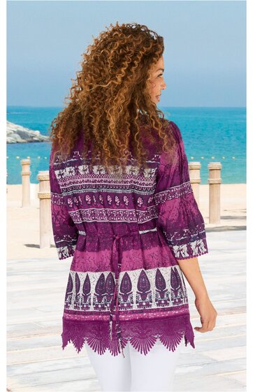 Lila und weiße Tunika mit floralem und geometrischem Muster, dreiviertellange Ärmel, leicht tailliert mit Gürtel, Spitzenabschluss am Saum, getragen von stehender Frau mit lockigen Haaren, Meereskulisse im Hintergrund