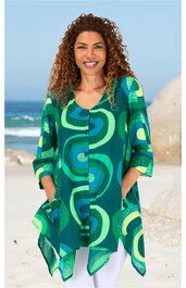 Locker geschnittene Bluse in Grün mit abstraktem Muster aus hellgrünen, blauen und türkisen Ovalen, V-Ausschnitt, durchgehende Knopfleiste, dreiviertel Ärmel, asymmetrischer Saum, getragen von stehender Frau mit gefalteten Händen