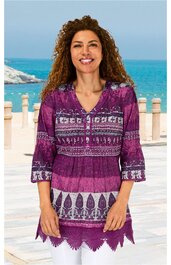 Lila Tunika mit weißen floralen Mustern, V-Ausschnitt mit Knopfleiste, 3/4-Ärmel mit Zierborte, gehäkelter Saum, getragen von stehender Frau vor Meereshintergrund