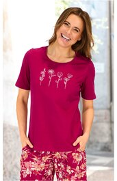 Dunkelrosa T-Shirt mit kurzen Ärmeln, Rundhalsausschnitt, weiß rosa Blumenmuster auf der Brust, getragen von lächelnder stehender Frau, kombiniert mit dunkelrosa Hose mit floralem Muster