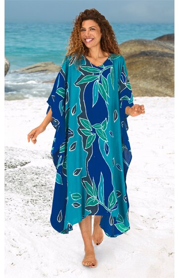 Kaftan in dunkelblau und türkis mit großflächigem floralem Muster in grün und hellblau, V-Ausschnitt, weite Form, knöchellang, getragen von lächelnder stehender Frau am Strand