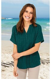 Locker geschnittene Bluse in dunklem Tannengrün mit feinem Karomuster, kurzer überschnittener Ärmel, V-Ausschnitt mit kleiner Knopfleiste, getragen von lächelnder Frau am Strand