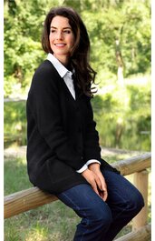 Schwarze Strickjacke mit Knöpfen, V-Ausschnitt, weiße Bluse mit Kragen darunter, dunkelblaue Jeans, sitzende Frau auf Holzgelaender im Freien, entspannte Pose