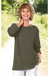 Olivgrüner Pullover ohne Muster, langer Schnitt, Rundhalsausschnitt, lange Ärmel, getragen von lächelnder stehender Frau mit kurzen blonden Haaren, eine Hand am Hals, eine in der Hosentasche